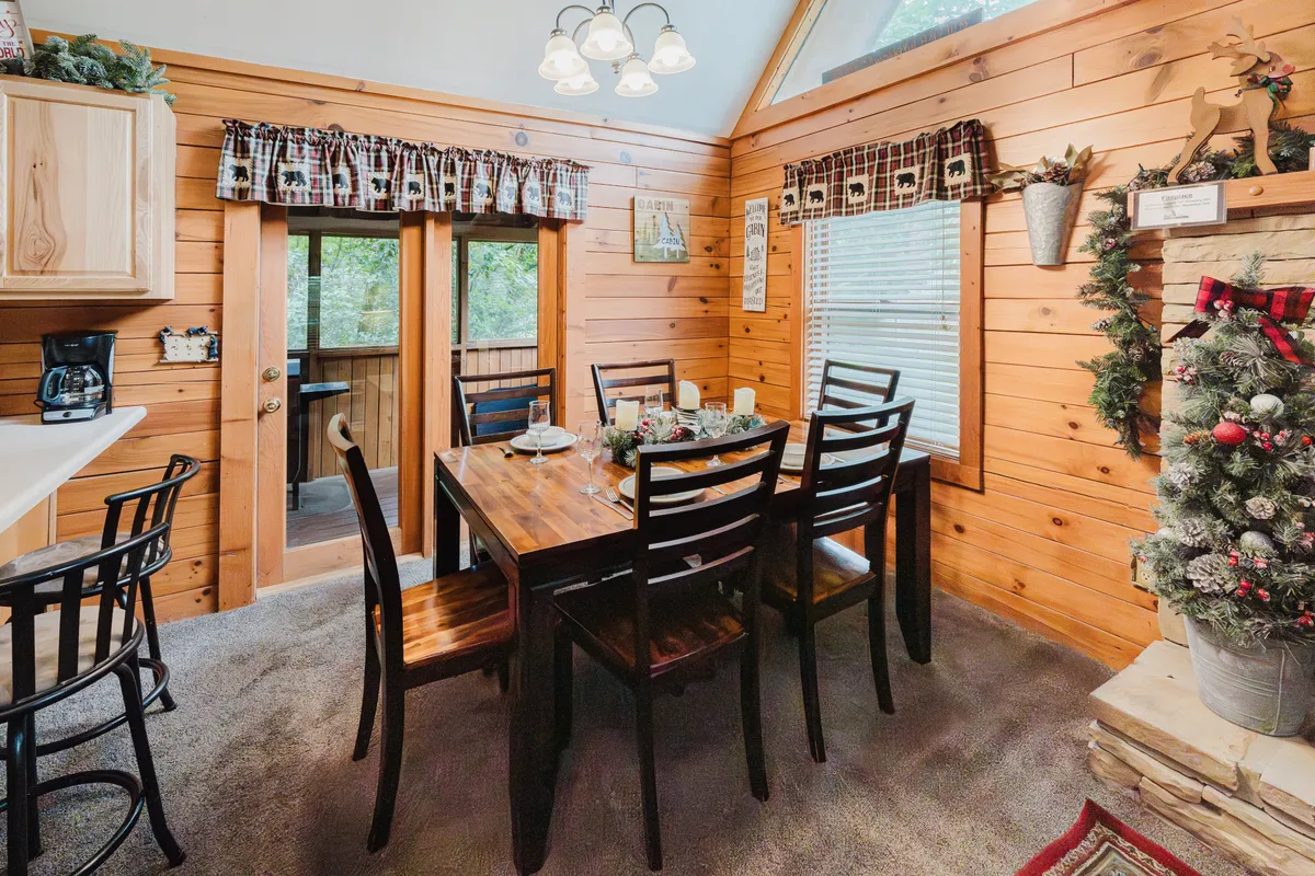 A large wooden dining table sits beside a glowing Christmas tree and expansive windows, offering a welcoming setting for family dinners, holiday celebrations, or friendly game nights. The open, airy space is decorated for the season, with the natural wood and festive accents enhancing the cozy atmosphere.