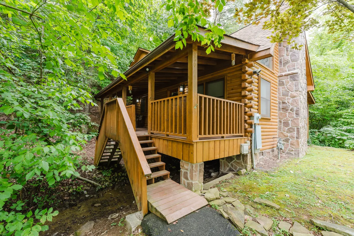 This image features the exterior of the log cabin, with inviting wooden steps, a solid stone foundation, and tranquil surrounding greenery. The porch is visible, suggesting a peaceful spot for morning coffee or evening relaxation, while the backdrop hints at the yard's expanse and the serenity of the wooded setting.