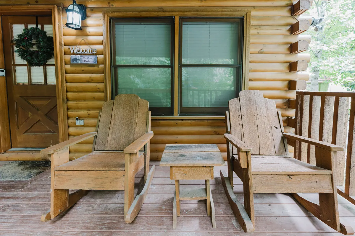 A cozy front porch with classic rocking chairs and a small table, dressed with a festive wreath on the wooden door. The porch invites guests to relax and enjoy the holiday spirit outside, surrounded by nature and rustic charm.