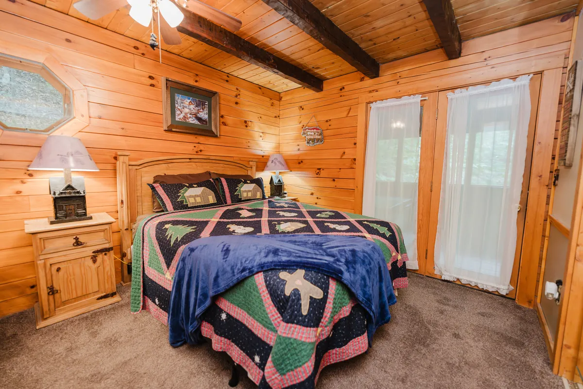 A cozy bedroom with a quilted bed, nightstands with lamps, wooden walls, and windows with curtains. The space feels warm and private, perfect for rest and relaxation.