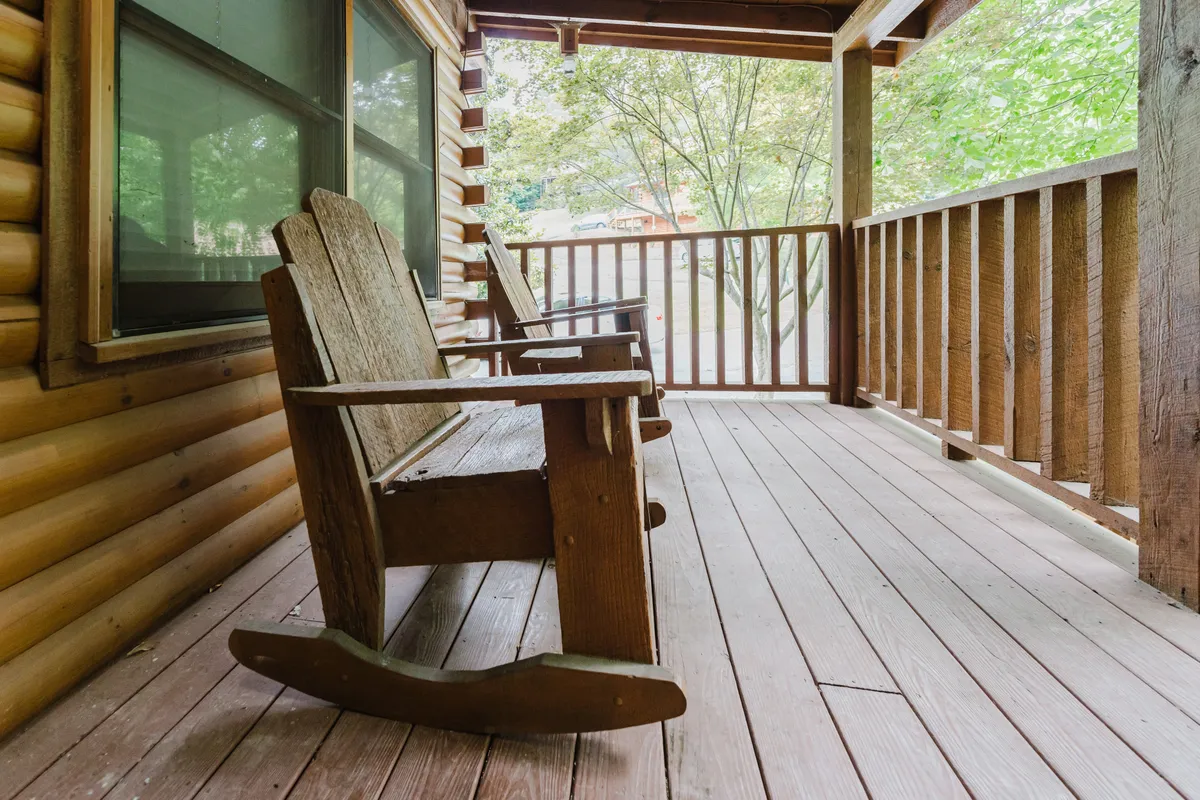 A cozy exterior view of a porch with rocking chairs and a wooden railing, surrounded by natural greenery. The scene evokes quiet moments spent sipping coffee, reading, or enjoying conversations against a tranquil backdrop.
