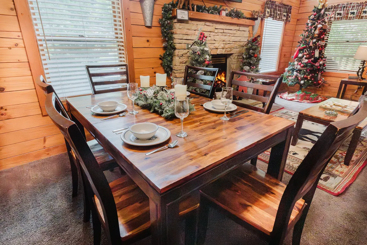 A warmly lit interior space featuring a wooden dining table set for an intimate meal, a glowing fireplace, and a decorated Christmas tree in the background. The scene exudes a festive, cozy atmosphere perfect for couples seeking a romantic getaway. The ambiance is inviting, with the fire adding an element of snuggly warmth ideal for relaxation and togetherness.
