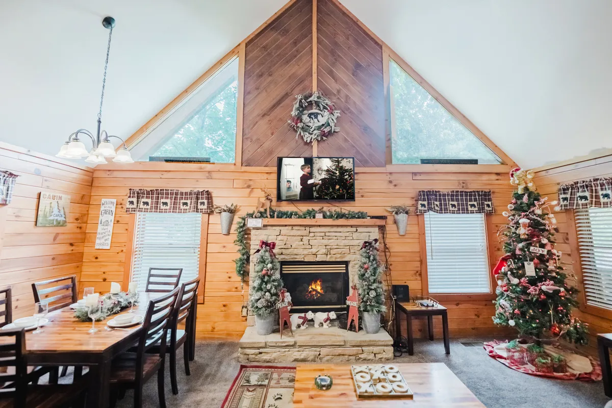 A beautifully decorated dining area with a wooden table, surrounded by warm lighting and accented by a glowing fireplace and a sparkling Christmas tree. The setup highlights holiday-themed décor, creating a scene of togetherness and seasonal celebration.