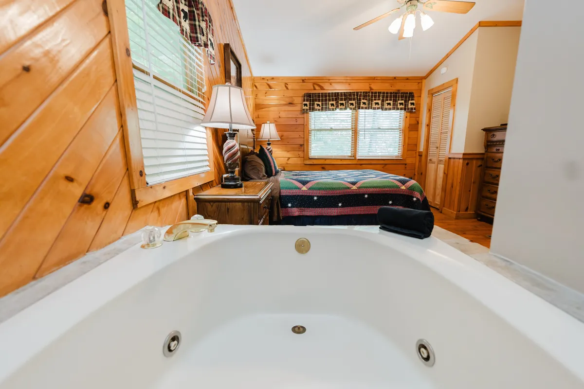 A loft-style bedroom featuring a wooden interior, a queen bed, a relaxing in-room Jacuzzi tub by the window, and soft lighting from a nearby lamp. The atmosphere is open and inviting, ideal for unwinding.