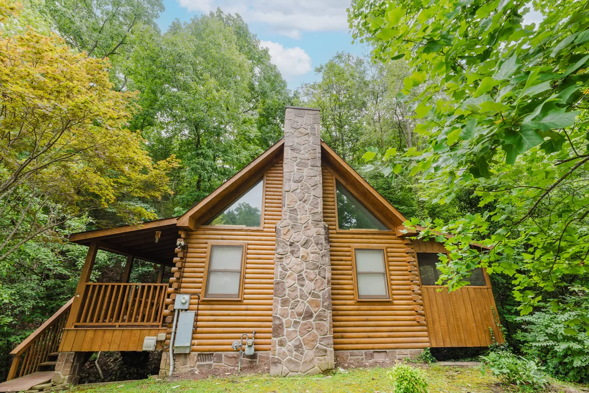 A wide view of the log cabin exterior, featuring a stone chimney, wooden deck, and lush green surroundings. The tranquil setting evokes classic Smoky Mountain charm and hints at the magical experiences awaiting inside and out.