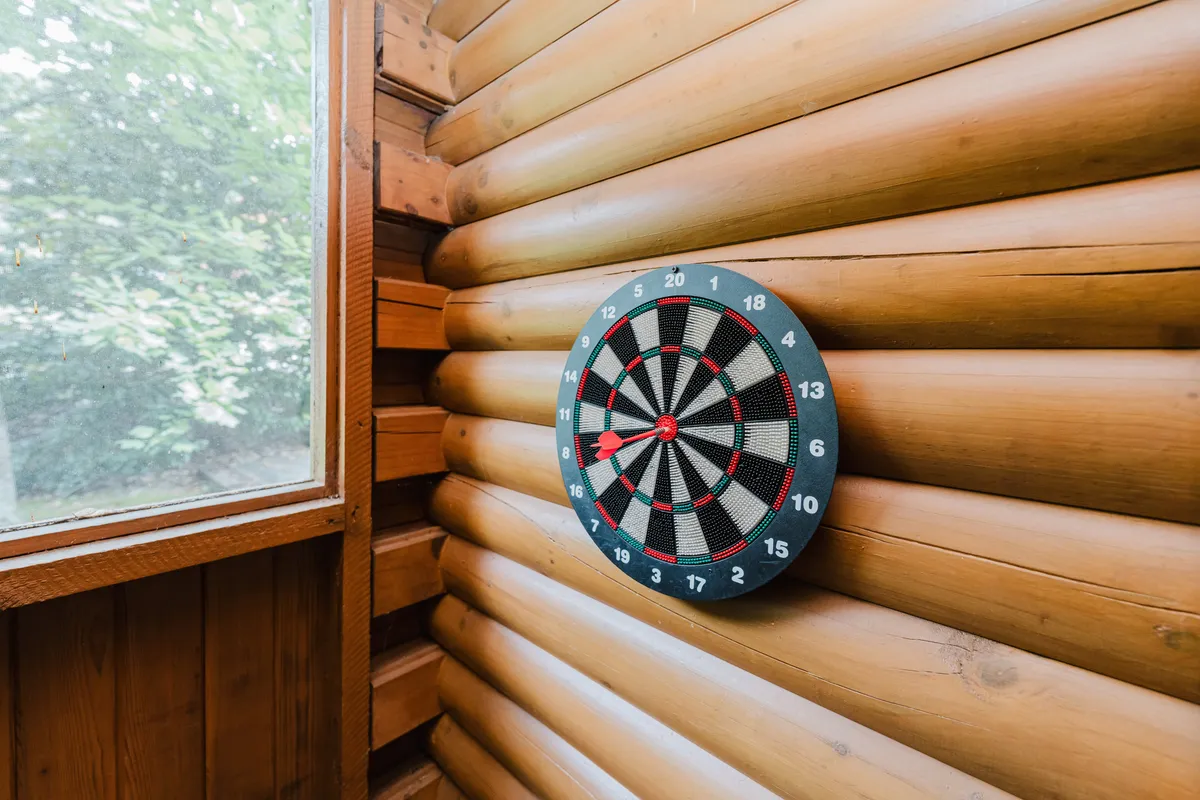 A playful corner featuring a dartboard on log walls with a window view of the outdoors, suggesting opportunities for fun, games, and bonding after a day of exploration.